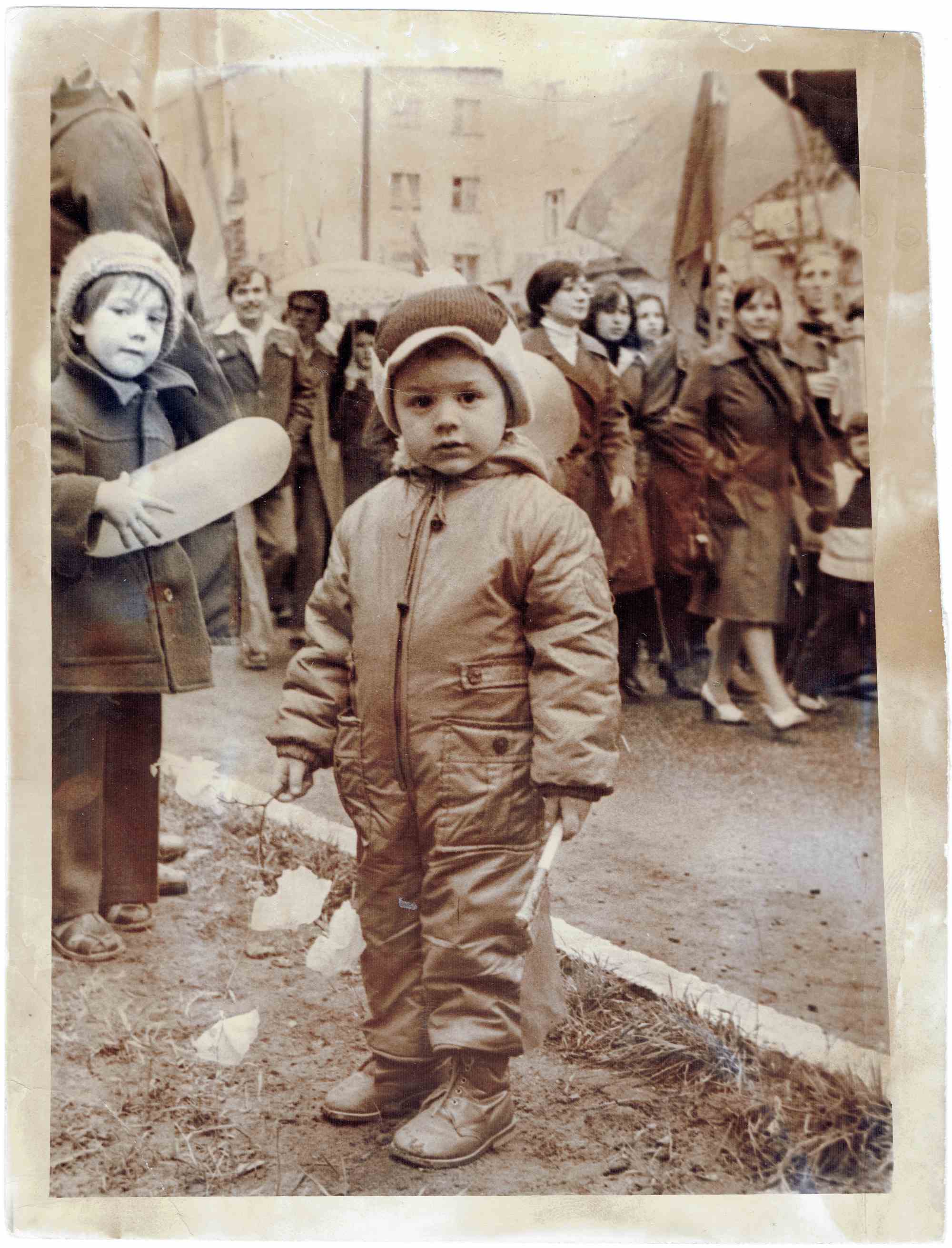 Сепия-фото: ребёнок в зимнем комбинезоне и шапке с ушами стоит на улице, держит флаг; рядом другой ребёнок с бумажным предметом, на заднем плане — люди и здания.