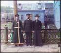 Три человека в тёмной одежде стоят на крыльце перед синим домом с деревянным забором.