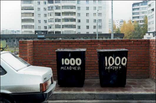 Два чёрных мусорных контейнера с надписью «1000 мелочей» на фоне кирпичной стены и жилых домов; слева видна часть белого автомобиля.