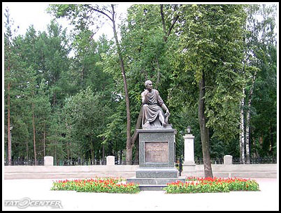 Бронзовая статуя сидящего мужчины на постаменте в парке, окружённая деревьями и цветочными клумбами с красными тюльпанами.