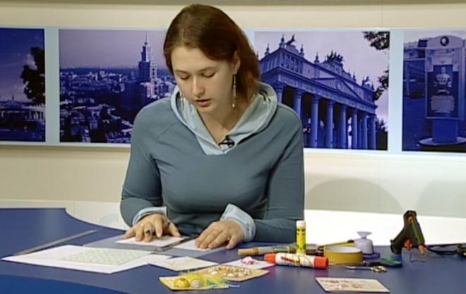 Женщина в сером худи сидит за столом с канцелярскими принадлежностями, работает с бумагами на фоне панорамных фотографий городских пейзажей и архитектуры.