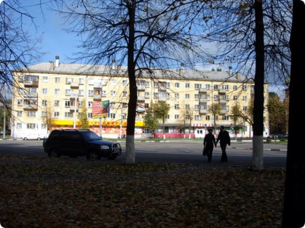 Два человека идут по осеннему парку мимо жёлтого многоэтажного дома с магазинами на первом этаже; на переднем плане — деревья и упавшие листья, на дороге — чёрный внедорожник.