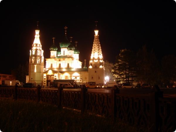 Ночная подсветка православного храма с зелёными куполами и двумя высокими шатровыми колокольнями, вид через тёмную кованую ограду на переднем плане.