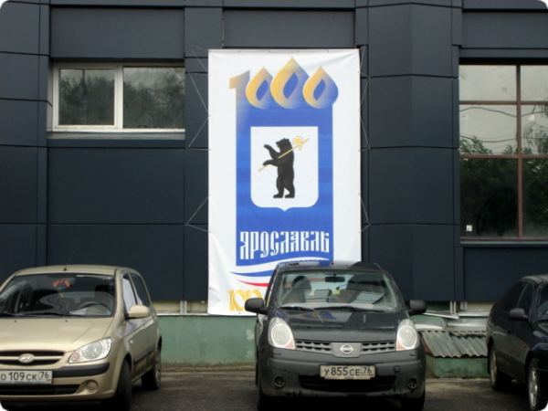 Баннер с гербом Ярославля (медведь с копьем) и надписью «Ярославль» на фасаде здания, перед ним припаркованы автомобили.