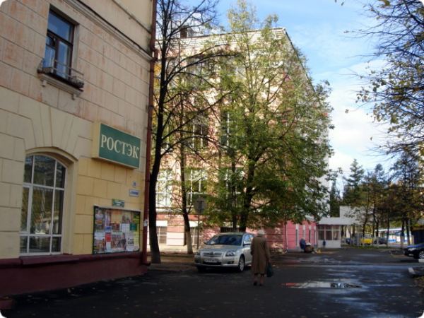 Жёлтое здание с вывеской «РОСТЭК» и информационным стендом, рядом — серебристый автомобиль, пешеход и деревья на фоне другого здания под облачным небом.