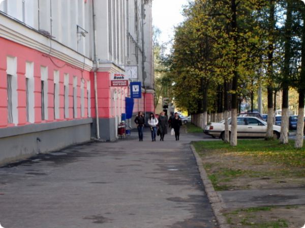 Четверо людей идут по тротуару вдоль здания с розово-белым фасадом и вывеской «Книжный магазин», рядом — ряд деревьев и припаркованные автомобили.