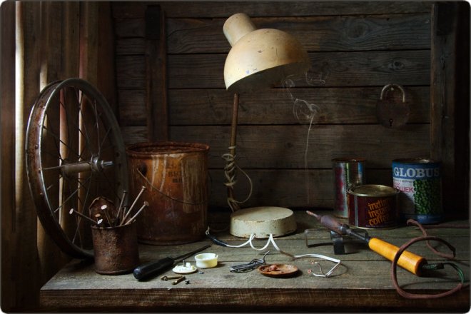 Ржавое велосипедное колесо, старая лампа, консервные банки, паяльник и инструменты на деревянном столе у стены.