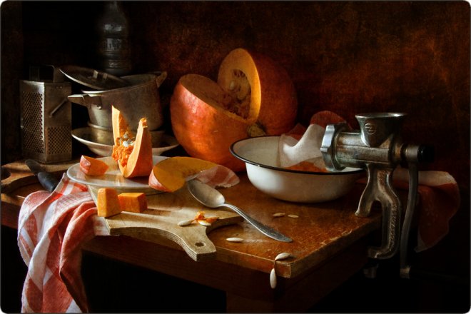 Натюрморт: разрезанная тыква, мясорубка, миска, доска с кусочками тыквы, ложка, полотенце и кухонная утварь на деревянном столе в тёплом свете.