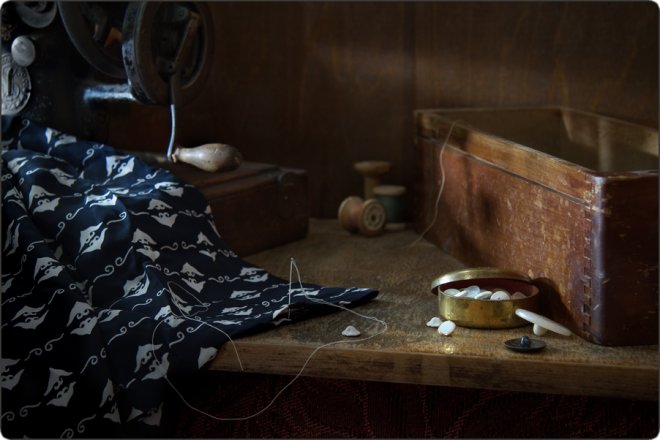 Старинная швейная машина на деревянном столе рядом с куском тёмной ткани с белым узором, катушками ниток, коробкой и открытой баночкой с пуговицами.