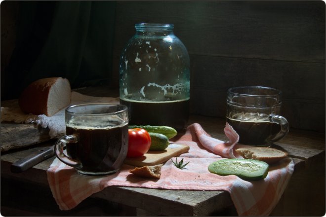 Светлая деревянная доска с ломтиком хлеба, помидором, огурцами, беконом, двумя стаканами тёмного напитка и стеклянной банкой на фоне тёмной стены.