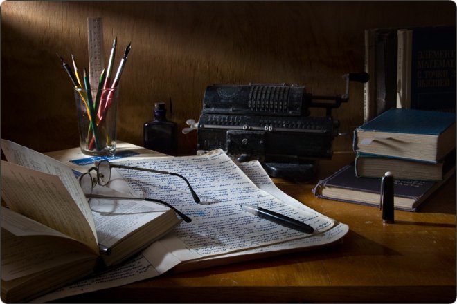 Рабочий стол с открытой книгой, рукописью, очками, пишущей машинкой, книгами и стаканом с карандашами в тёплом свете.