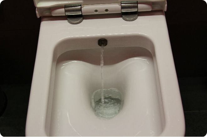 Белый подвесной унитаз с открытой крышкой, из которого течёт струя воды в чашу на тёмном фоне.