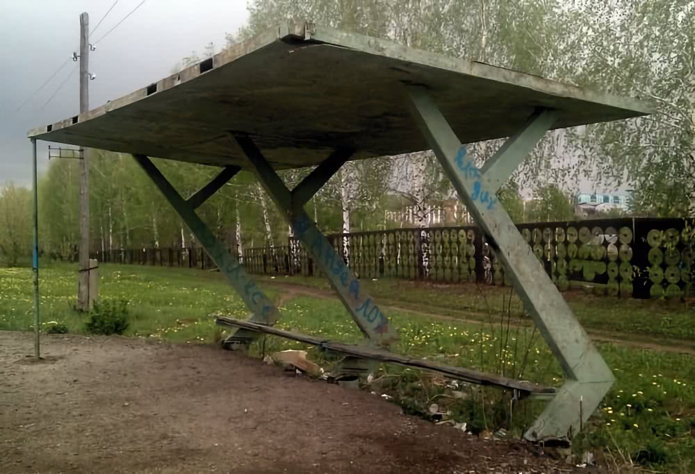 Заброшенный металлический павильон с полом и скамьей, покрытый граффити, стоит на обочине среди травы и берёз; на заднем плане виден забор и здание.