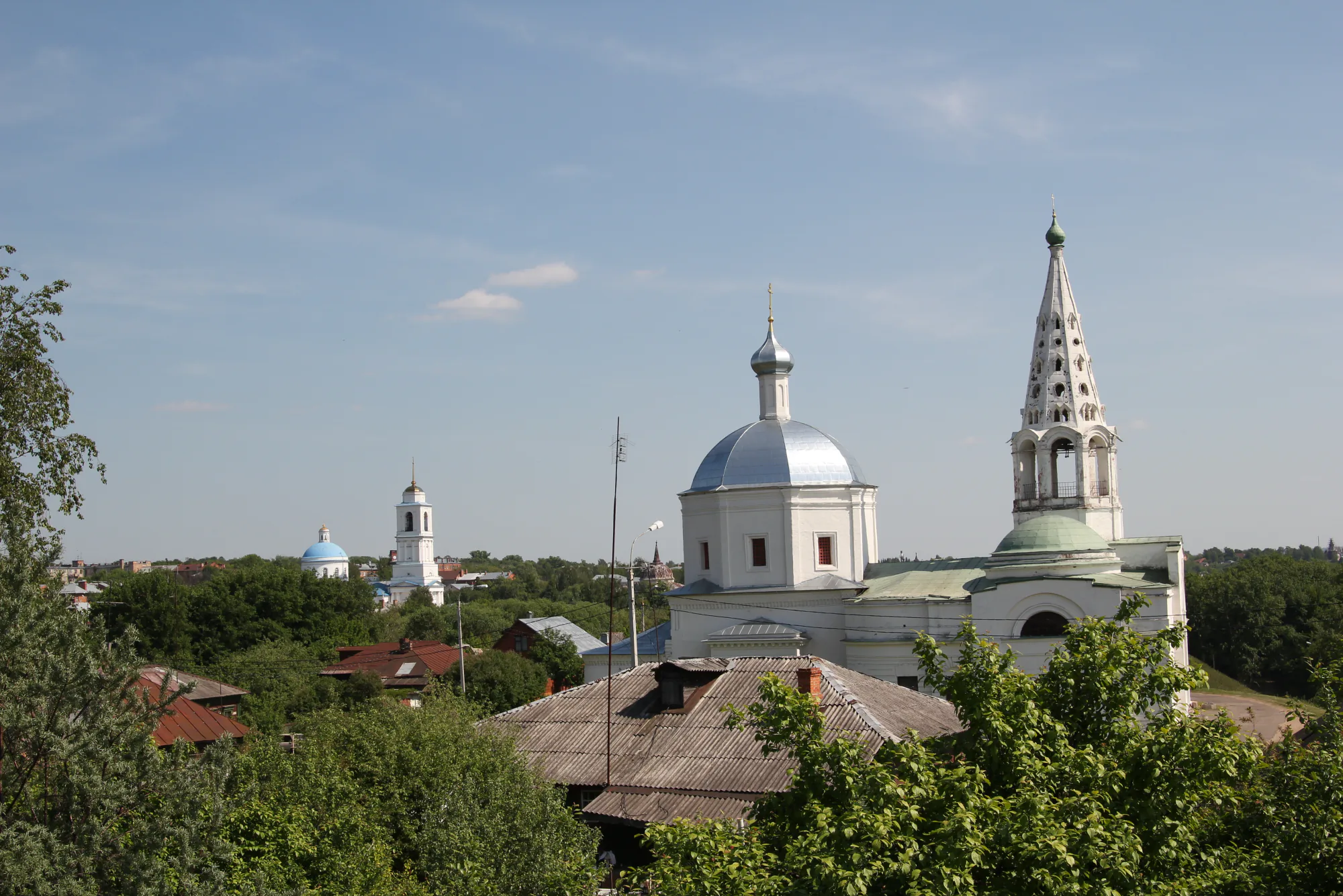 Белые храмы с куполами и колокольней на фоне голубого неба, видны крыши домов и зелёные деревья в переднем плане.