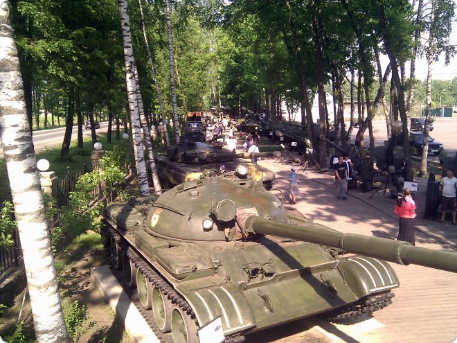 Военные танки выставлены на открытом воздухе в парке, посетители осматривают экспонаты среди берёз и дорожек.