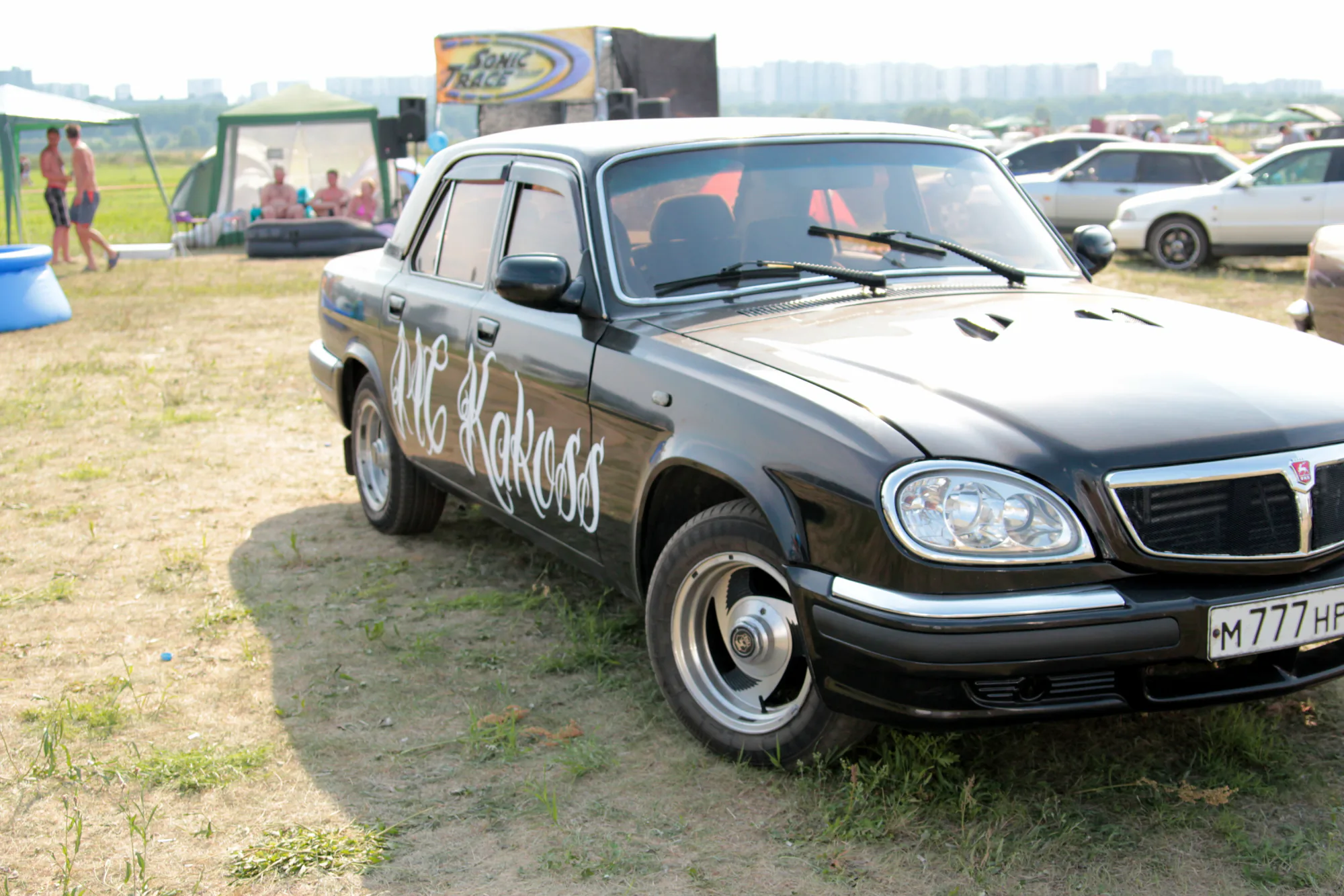 Чёрный автомобиль ГАЗ-3110 с надписью «MC KAKOSS» на двери припаркован на травянистом поле; на заднем плане видны палатки, люди и другие машины.