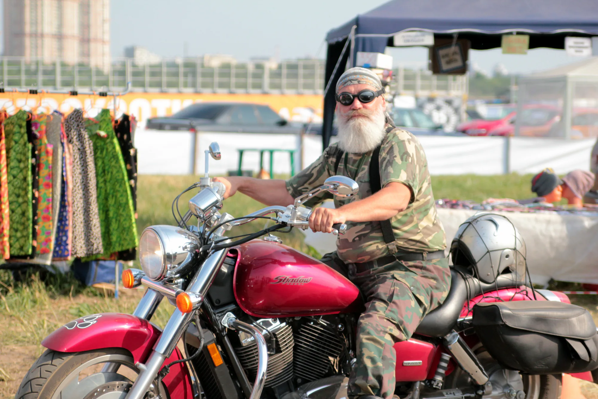 Мужчина в камуфляже и с белой бородой сидит на красном мотоцикле Honda Shadow на открытом воздухе, рядом с прилавком с тканями и палаткой.