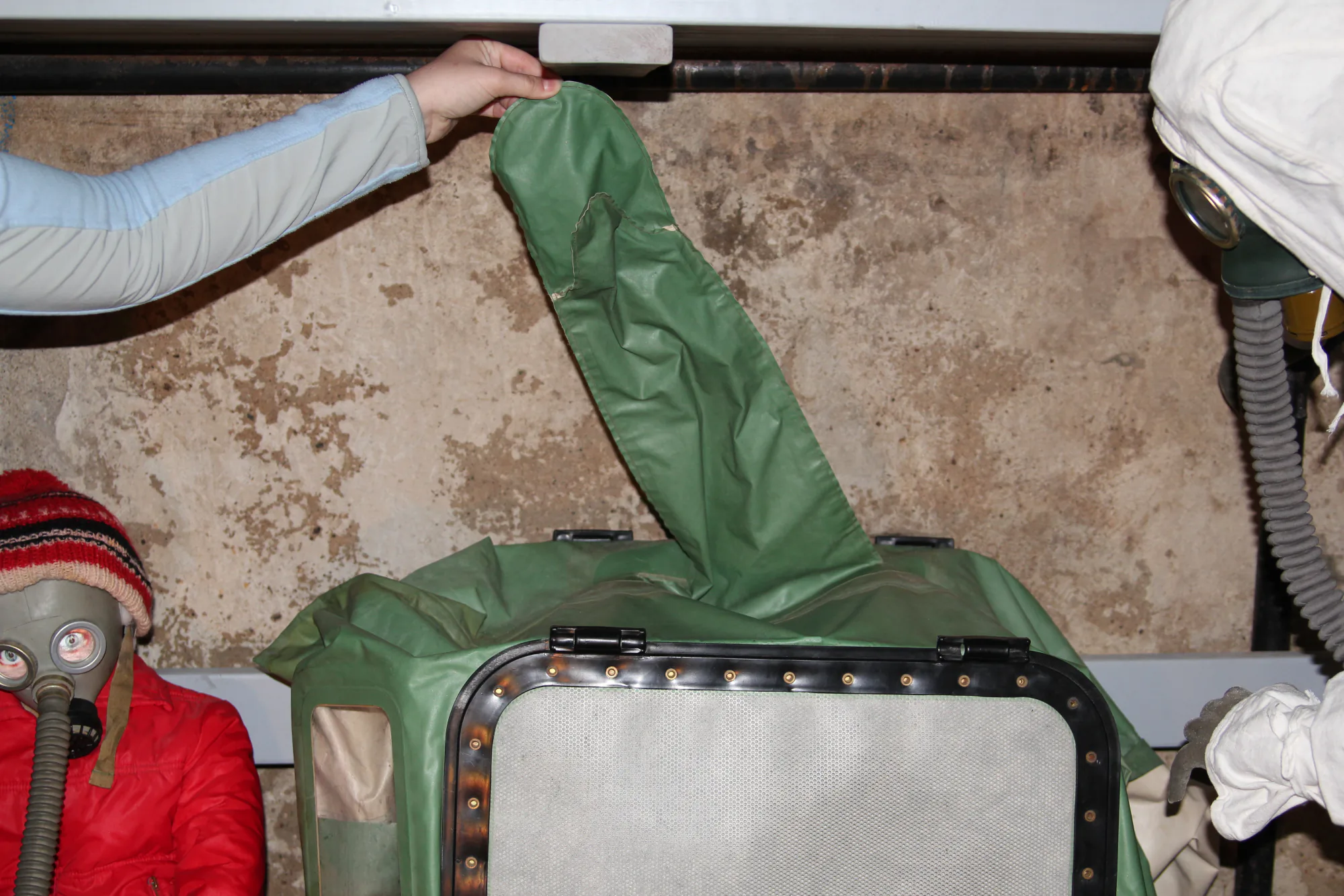 Рука в светло-синей перчатке держит зелёный тканевый рукав, присоединённый к защитному костюму с фильтром; рядом ребёнок в противогазе и красной куртке.