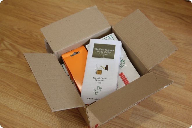 Открытая картонная коробка на деревянном полу, внутри — каталог «The Kent & Sussex Tea & Coffee Company» за 2017 год, оранжевая упаковка и другие товары.