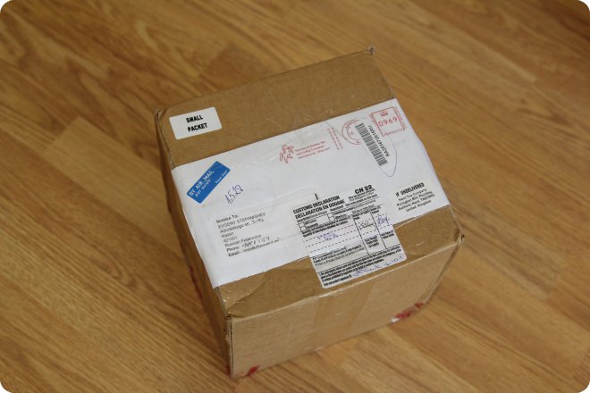 Картонная коробка с наклейкой «SMALL PACKET» и таможенной декларацией, лежащая на деревянном полу.