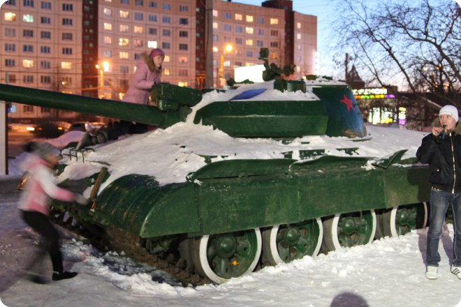 Зелёный танк Т-34, покрытый снегом, стоит на городской площади вечером; рядом играют дети, на башне видна красная звезда, на фоне — многоэтажный дом с освещёнными окнами.