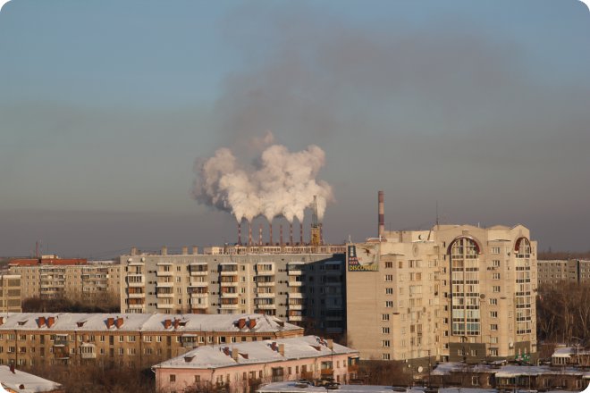 Зимний городской пейзаж: многоэтажные жилые дома с заснеженными крышами на переднем плане, на заднем плане — промышленные трубы, выбрасывающие густой белый дым в пасмурное небо.