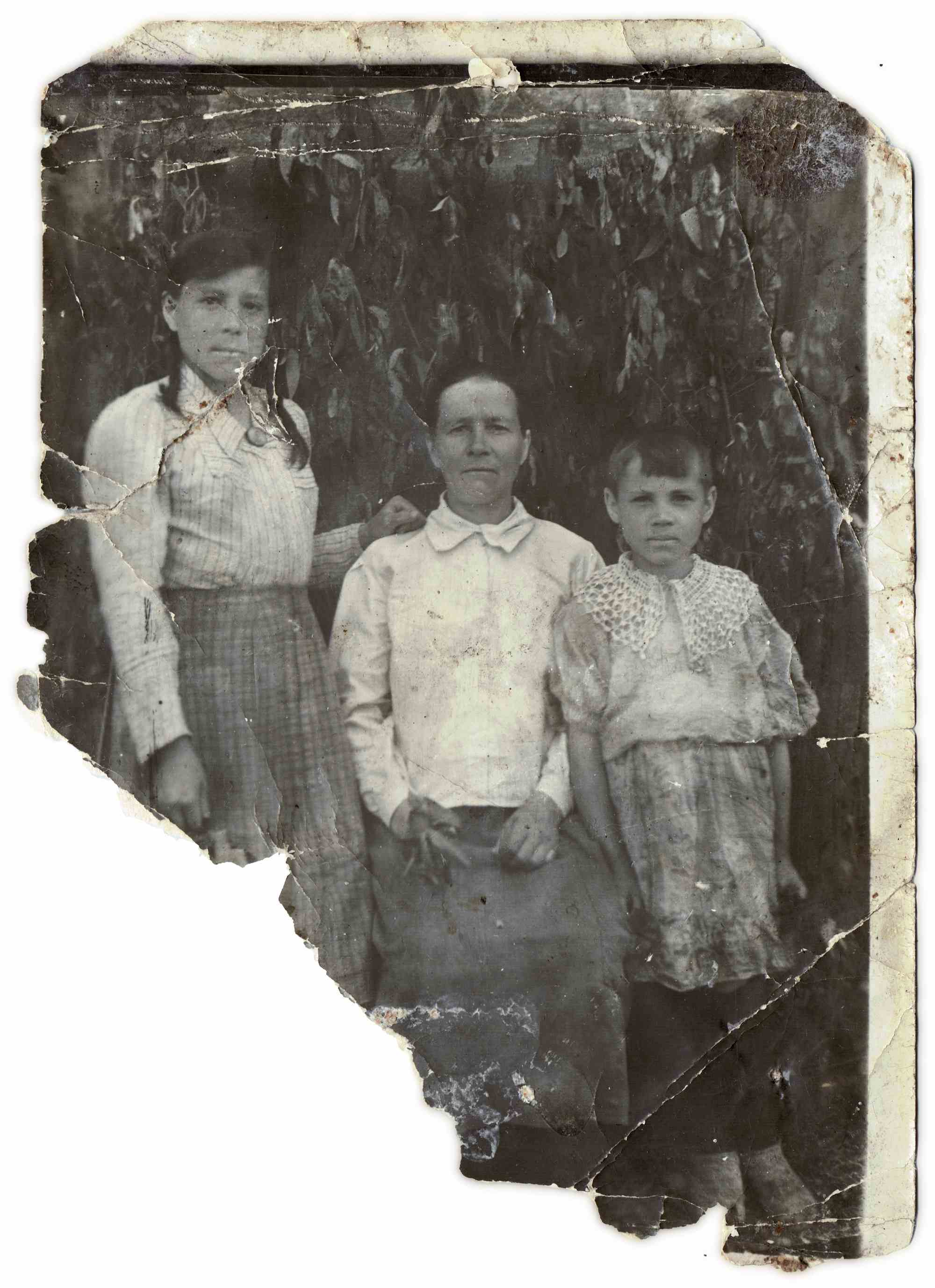 Потрёпанная чёрно-белая фотография: женщина сидит в центре, рядом с ней стоят девушка и девочка; фон — листва, видны трещины и потёртости.