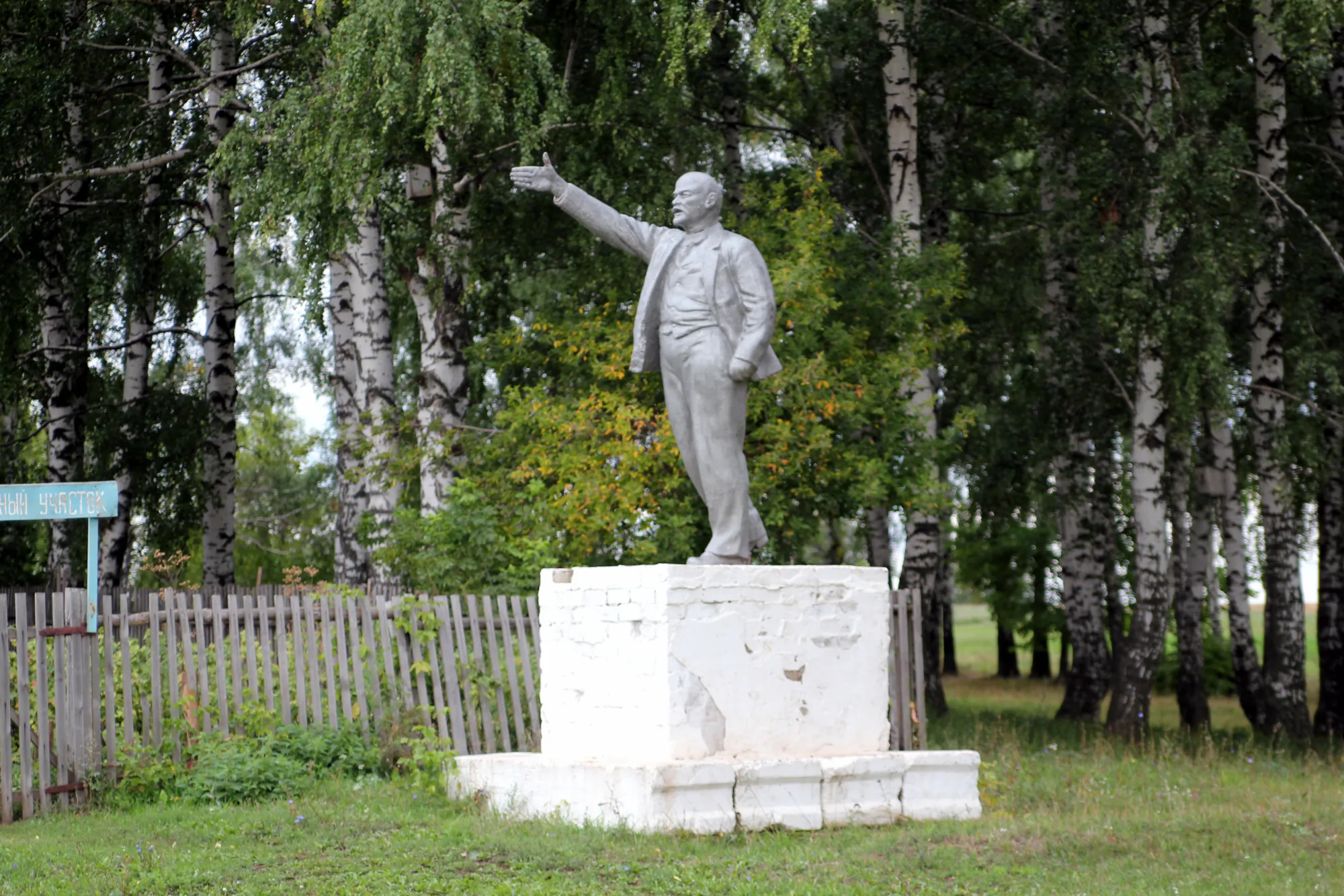 Белая статуя Ленина на постаменте в парке, мужчина в костюме стоит, протянув правую руку вперёд, на фоне берёз и деревянного забора с синей табличкой.
