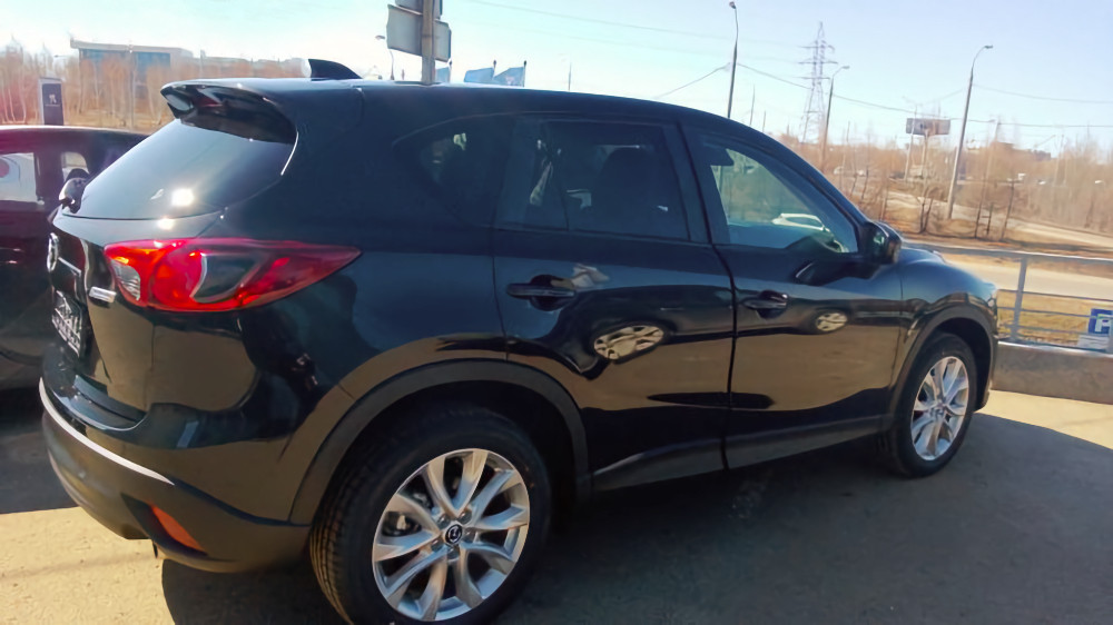 Чёрный кроссовер Mazda CX-5 на парковке, вид сзади-сбоку, солнечный день, на фоне дороги и промышленных зданий