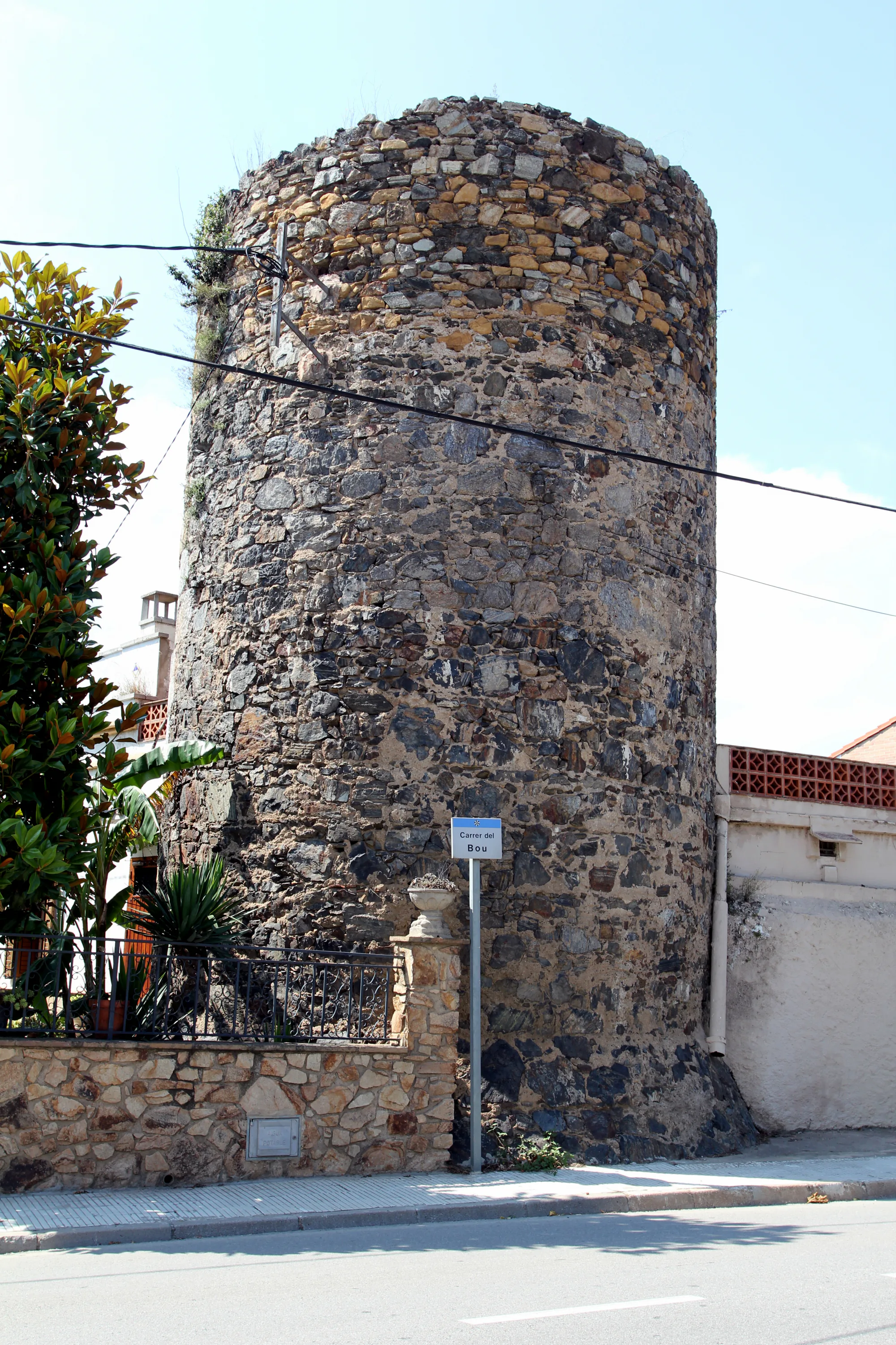 Старая круглая каменная башня на улице Carrer del Bou, окружённая зеленью и забором, под ясным небом.