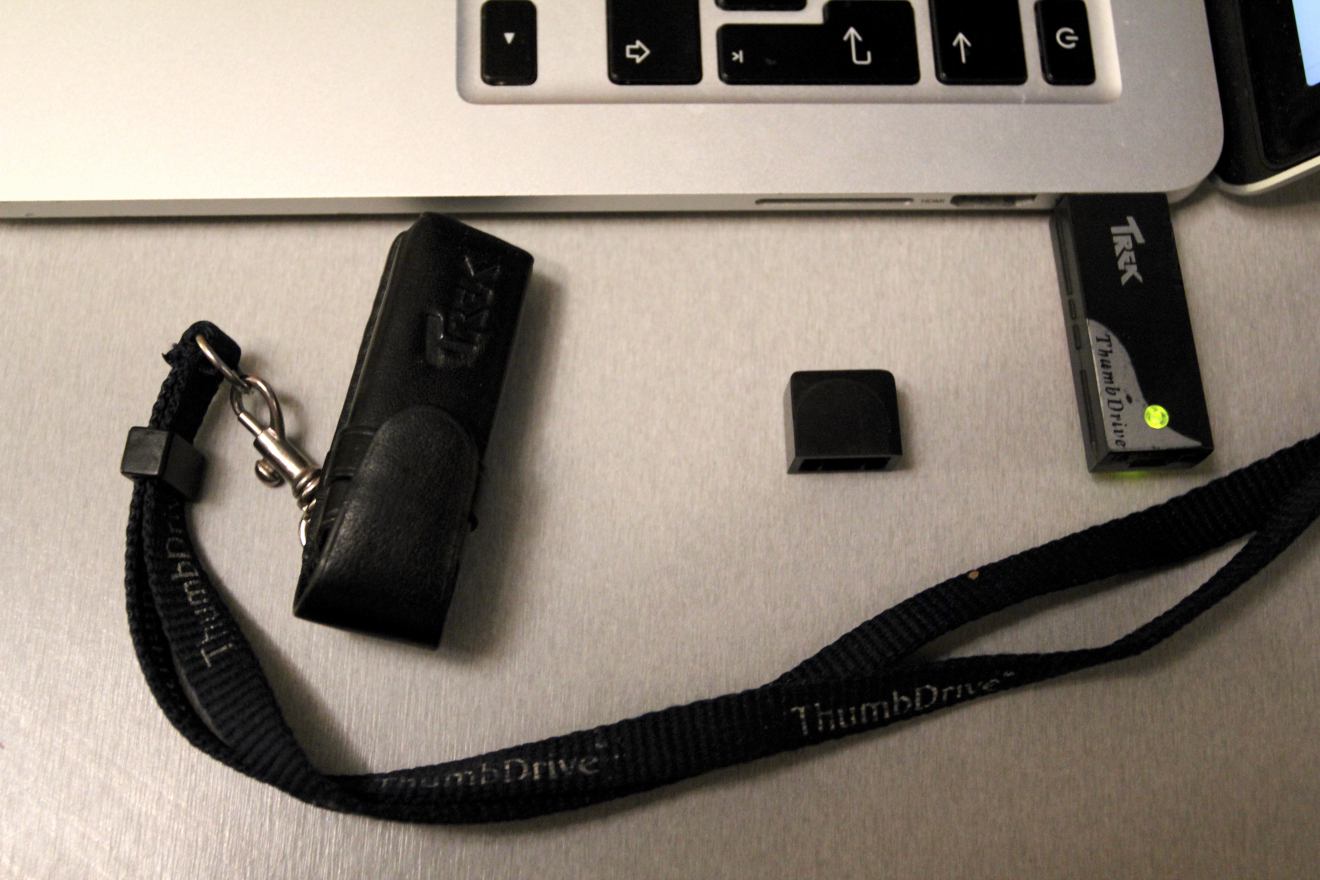 Чёрный USB-накопитель TREK ThumbDrive с зелёным индикатором подключён к ноутбуку; рядом — его кожаный чехол с логотипом и ремешком «ThumbDrive», а также отдельная чёрная крышка.