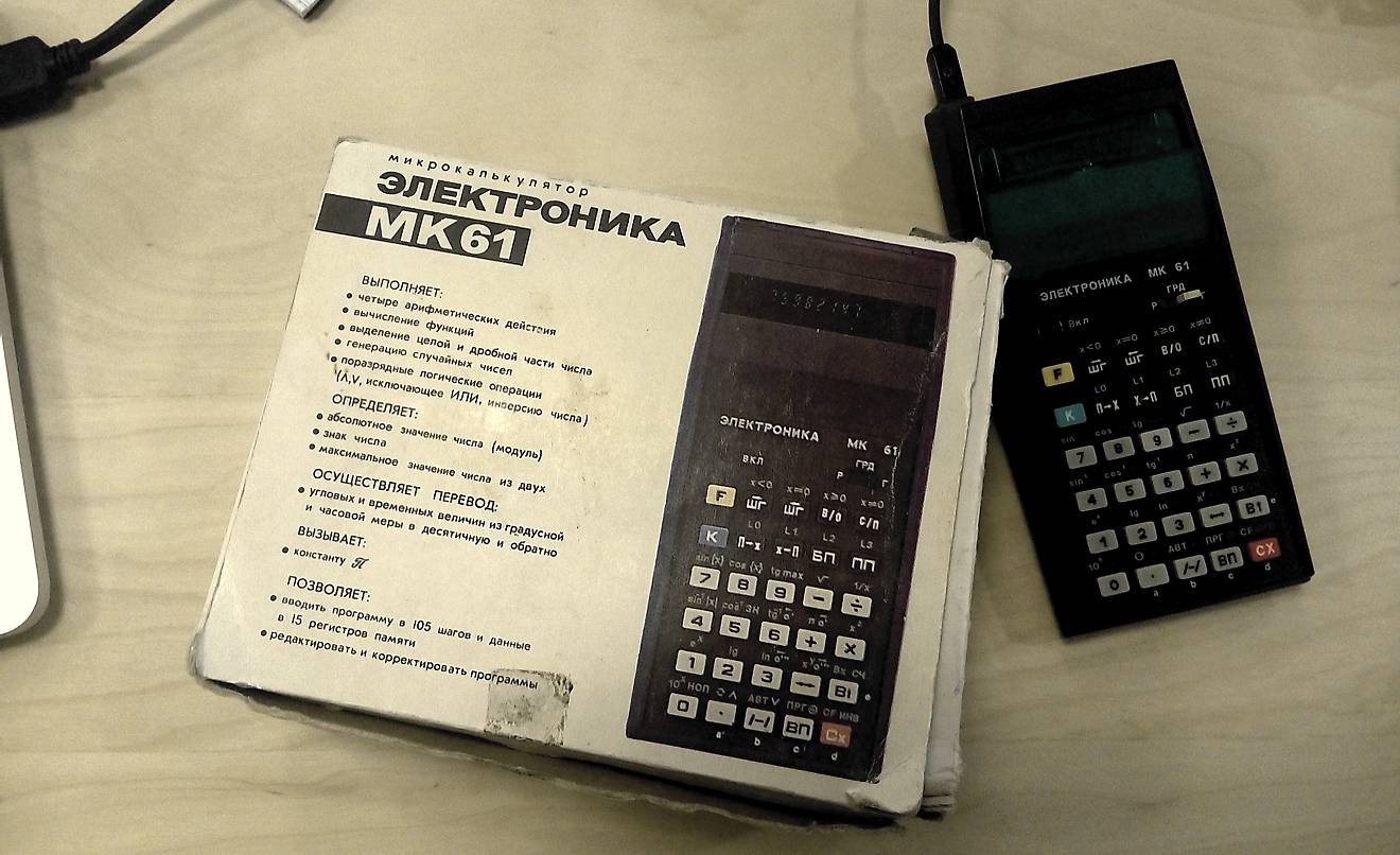 Микрокалькулятор Электроника МК-61 в картонной упаковке с описанием функций и рядом с самим устройством на светлом столе.