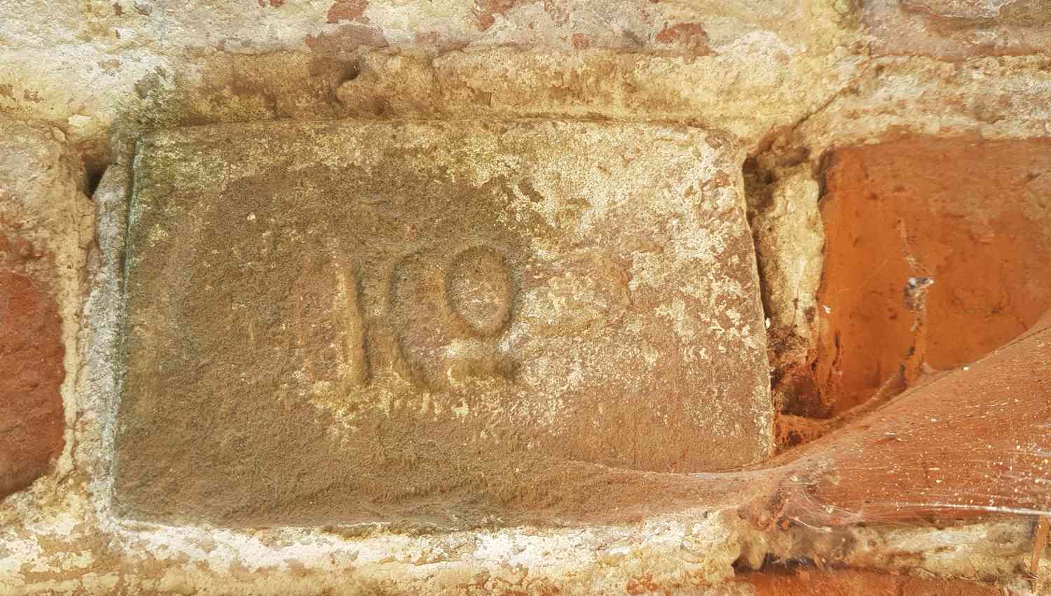 Старый кирпич с выгравированной цифрой «19», покрытый пылью и паутиной, вставленный в стену из красного кирпича и штукатурки.