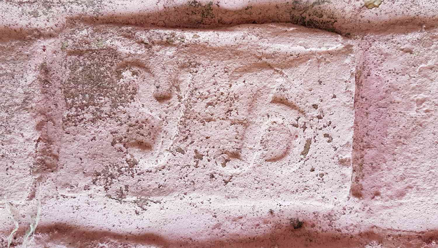 Выцветшая розовая кирпичная кладка с вырезанным на одном из кирпичей символом, напоминающим букву «С» или полумесяц, покрытая пылью и мхом.