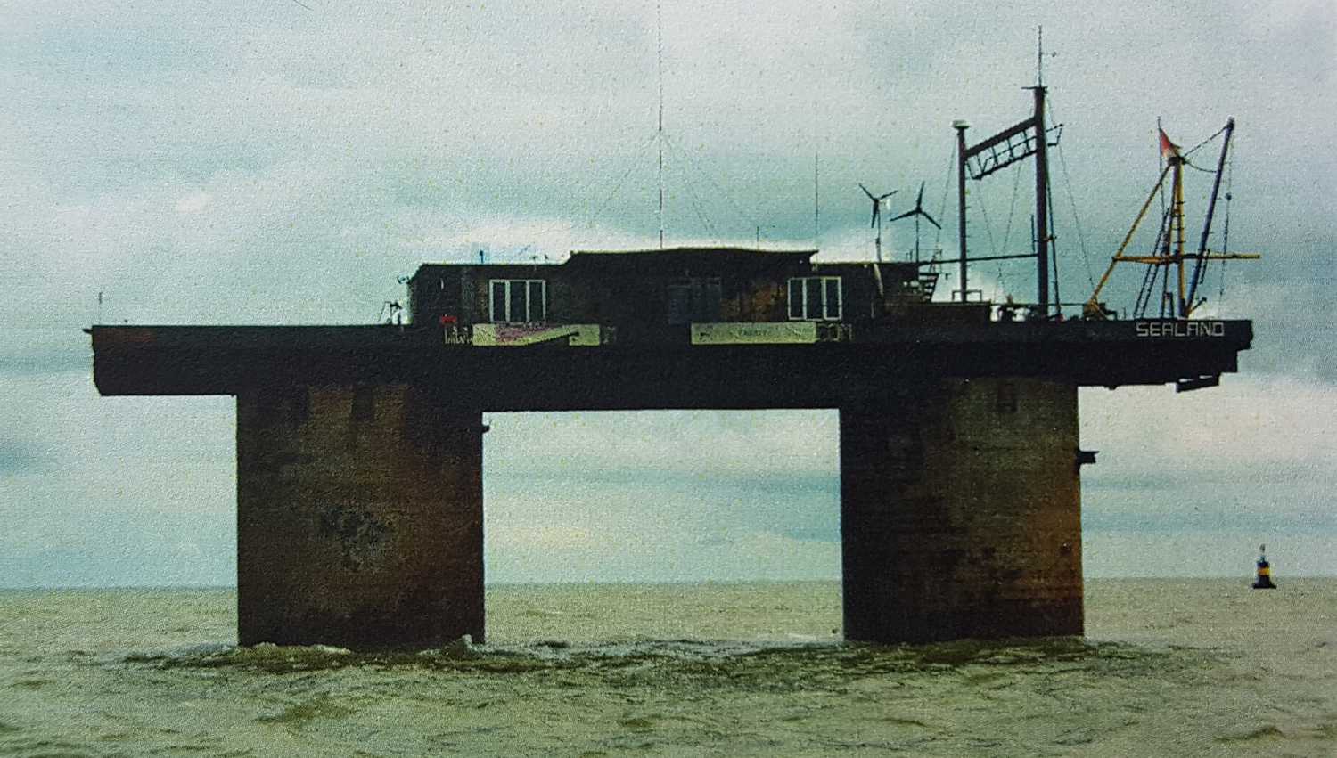 Морская платформа «Sealand» с жилыми модулями, антеннами и ветрогенераторами на фоне облачного неба и моря.
