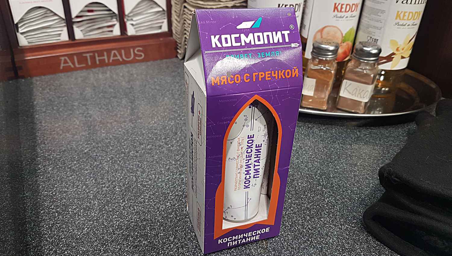 Фиолетовая упаковка «Космопит» с надписью «Мясо с гречкой» и «Космическое питание», внутри туба с аналогичной маркировкой, на столешнице рядом с банками специй.