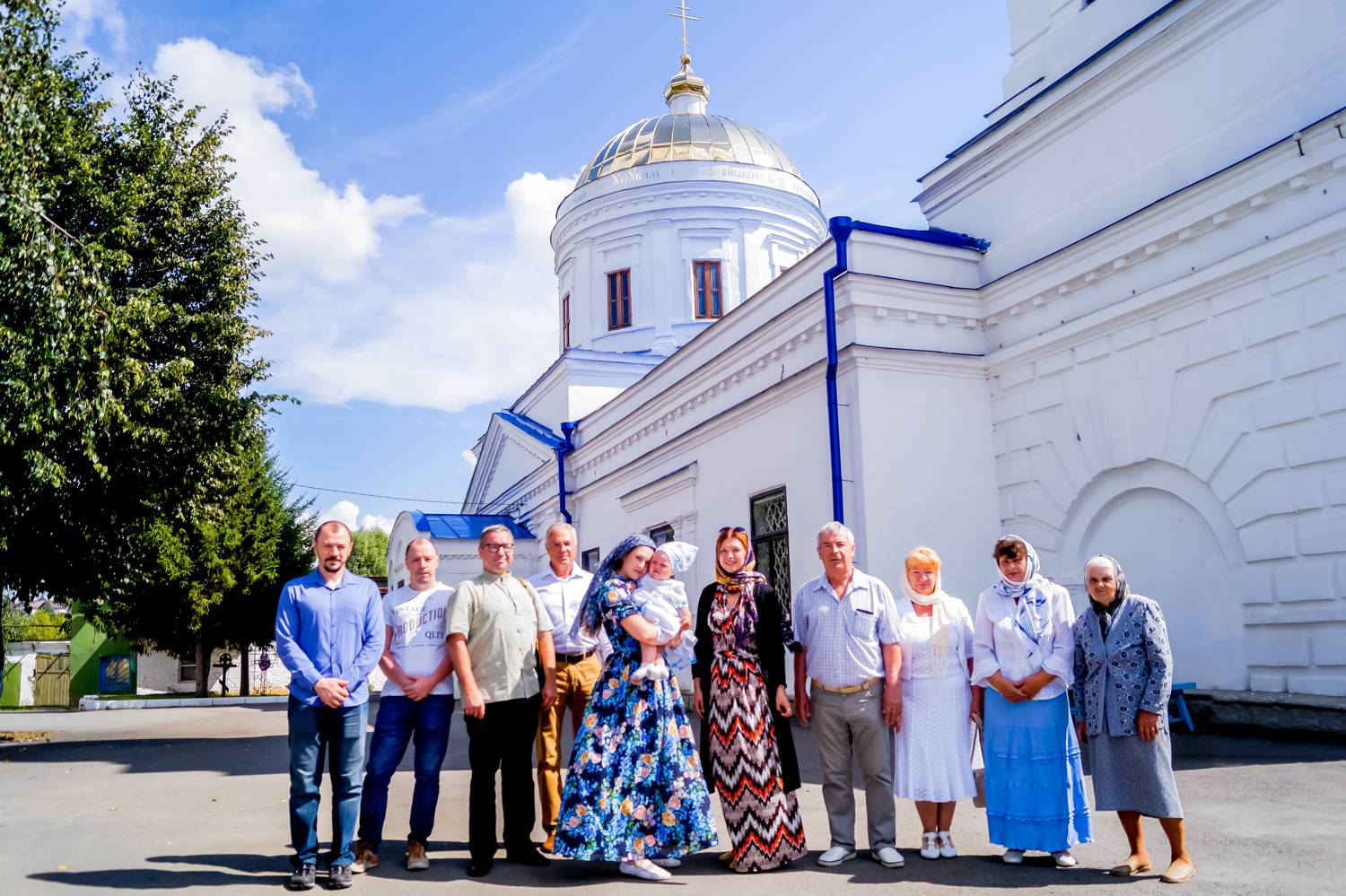 Группа из десяти человек, включая мужчину в рясе и женщину с младенцем, стоит на фоне белого храма с золотым куполом под голубым небом.