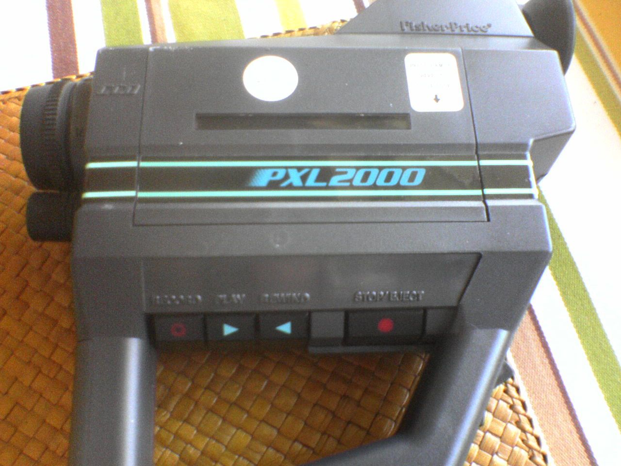 Черная игрушечная видеокамера Fisher-Price PXL2000 с кнопками управления и логотипом на корпусе, лежащая на плетеной поверхности.