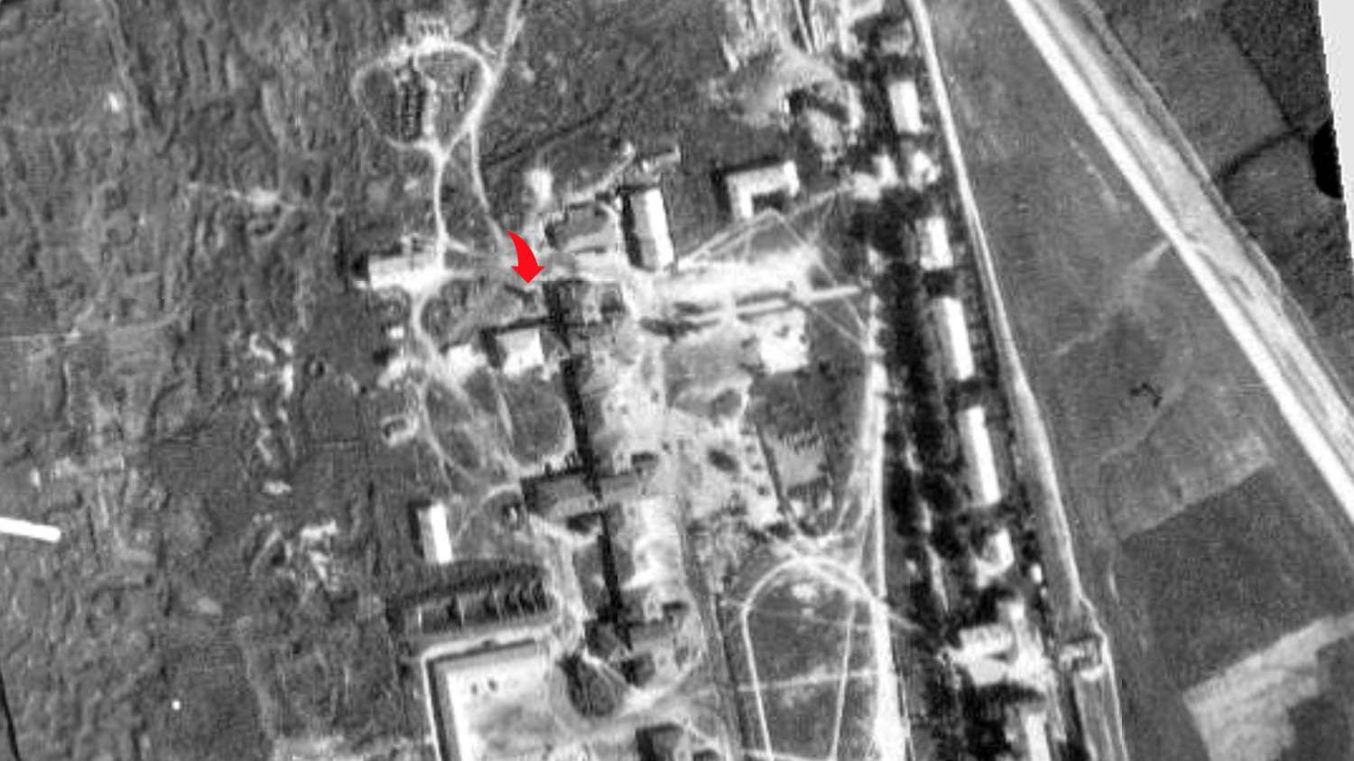 Аэрофотоснимок военного объекта с красной стрелкой, указывающей на одно из зданий в центре комплекса, окружённого дорогами и полями.