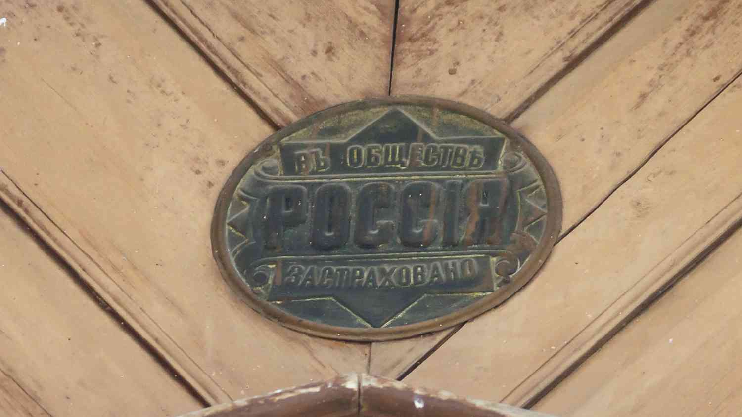 Металлическая овальная табличка на деревянной поверхности с надписью «ВЪ ОБЩЕСТВѢ РОССІЯ ЗАСТРАХОВАНО» старинным шрифтом.