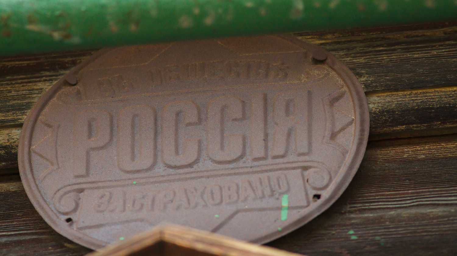 Ржавая овальная металлическая табличка с надписью «РОССИЯ» и «ЗАСТРАХОВАНО», прикреплённая к деревянной поверхности под зелёным элементом.