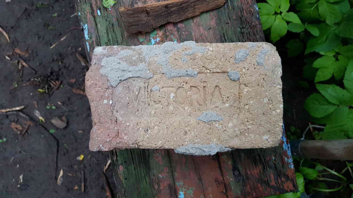 Старый кирпич с надписью «VICTORIA» и следами цемента лежит на потёртой деревянной поверхности среди земли и зелёных листьев.