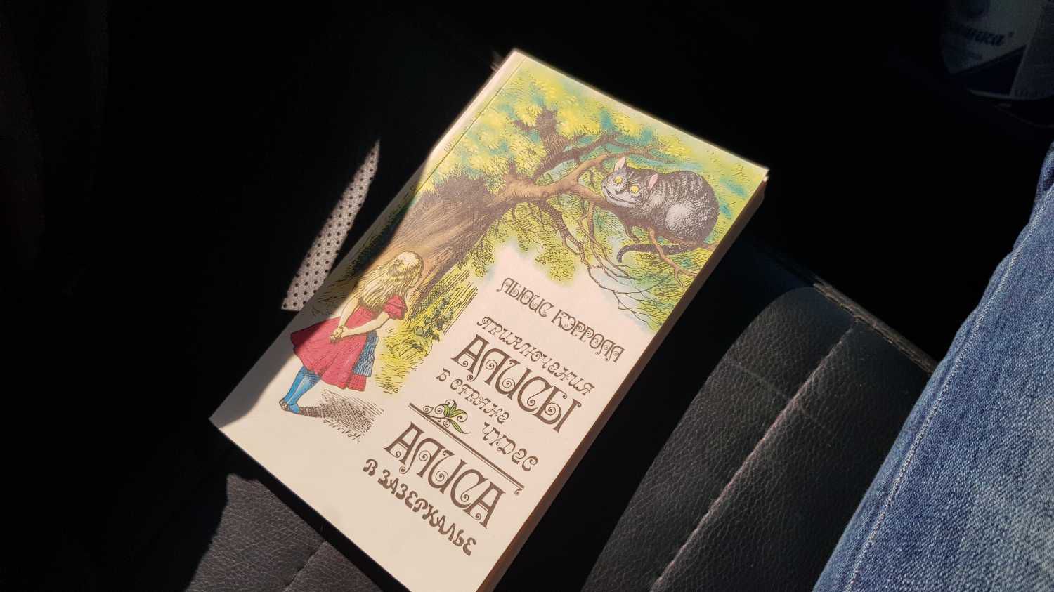 Обложка книги «Приключения Алисы в стране чудес» и «Алиса в Зазеркалье» Льюиса Кэрролла с иллюстрацией девочки и кота на дереве, лежащая на тёмном сиденье рядом с джинсами.
