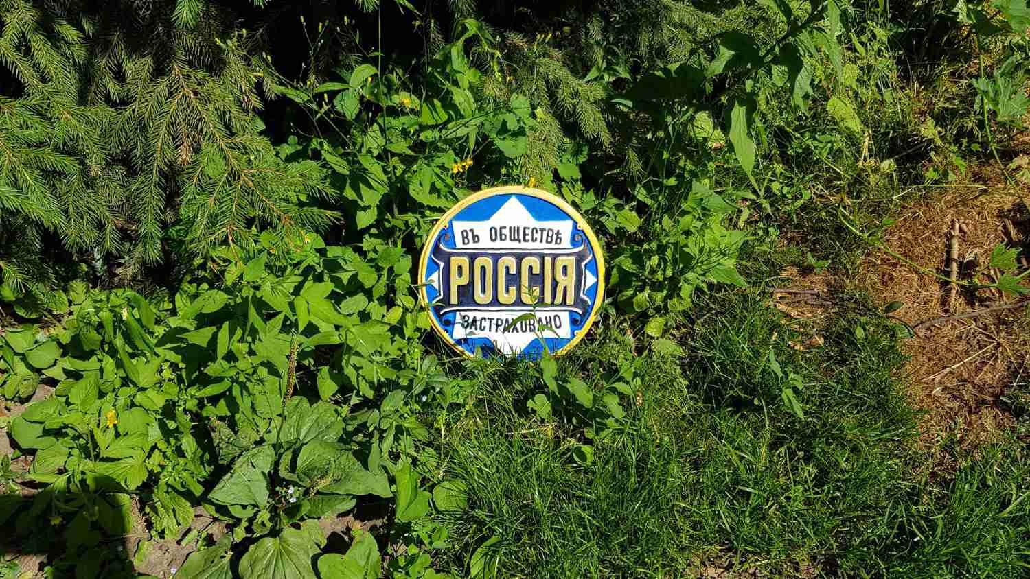 Круглая эмблема «Въ обществѣ РОССІЯ ЗАСТРАХОВАНО» на фоне зелёных листьев и хвои.