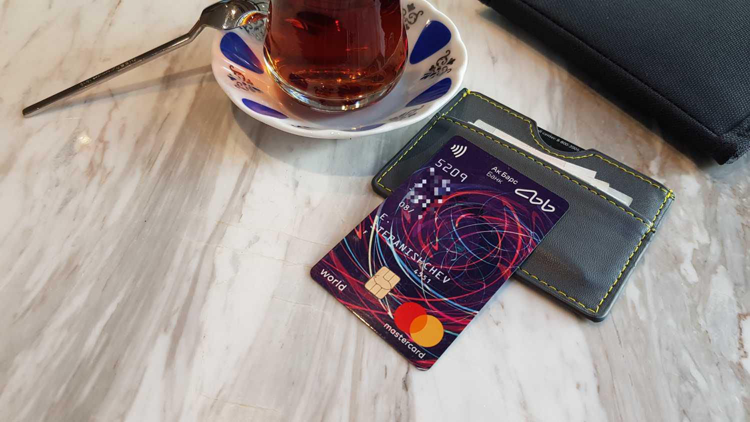 Банковская карта Mastercard World от Ак Барс Банка с именем владельца и номером, частично скрытым, лежит на сером кошельке с жёлтой строчкой рядом с чашкой чая на мраморном столе.