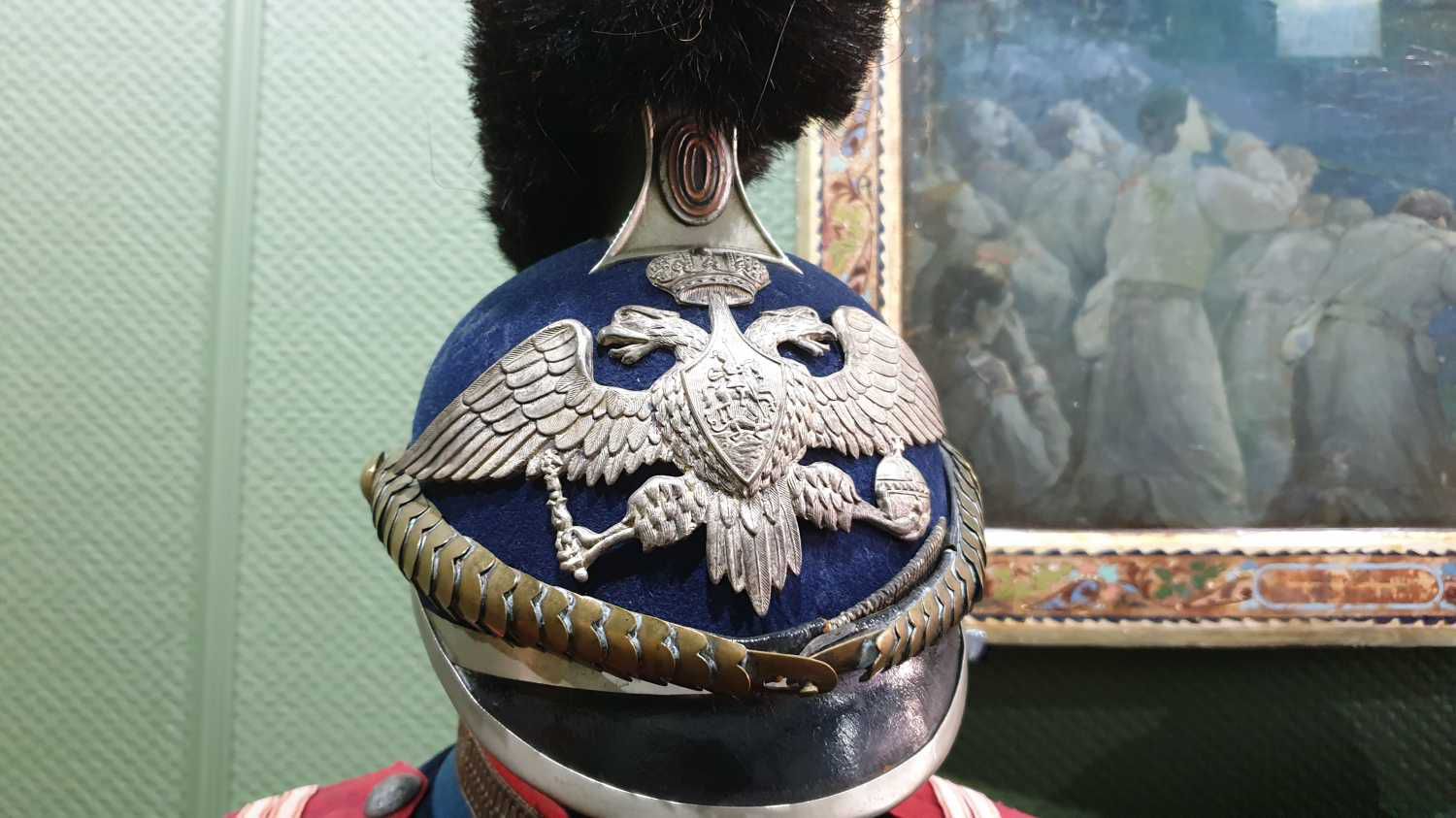 Закрытый шлем с двуглавым орлом и черным сулманом, выставленный в музее на фоне картины с исторической сценой.