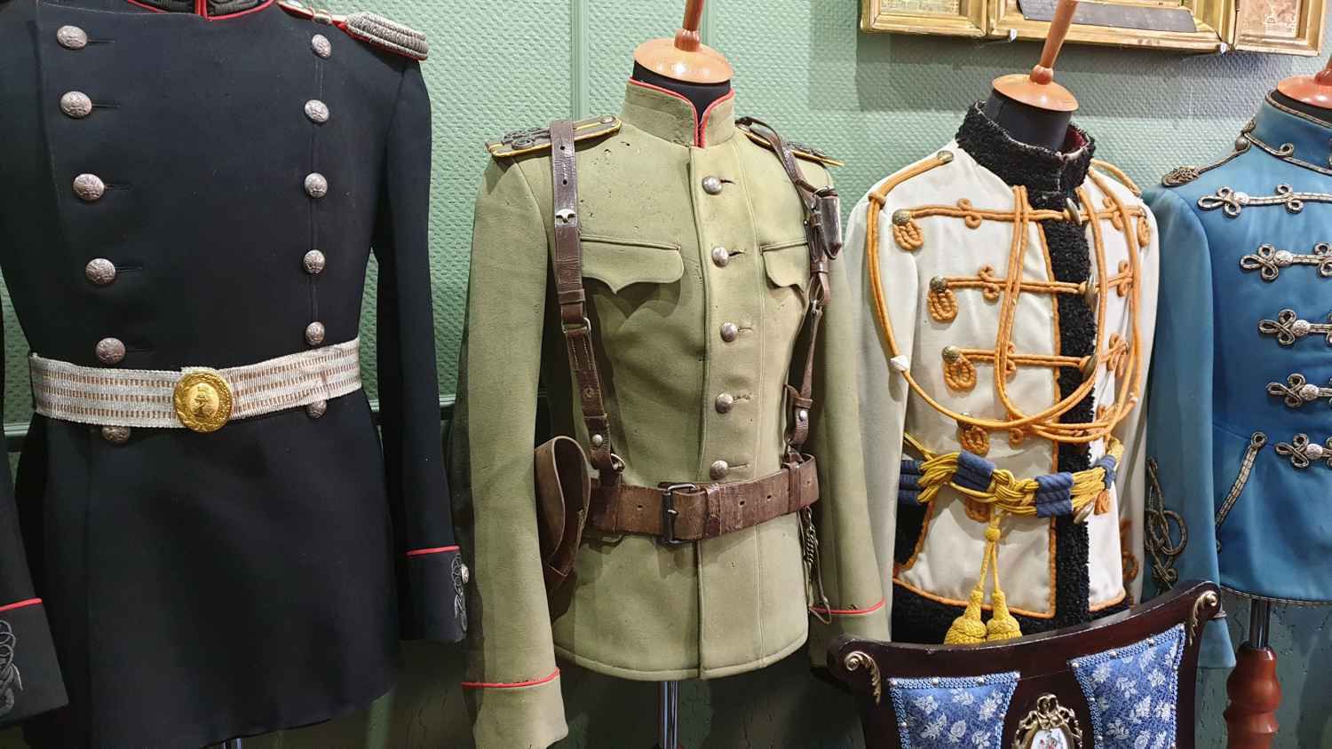 Четыре манекена в военной форме: чёрная с золотым ремнём, зелёная с кобурой, белая с золотым шнуром и синяя с серебряными пуговицами на фоне зелёной стены.