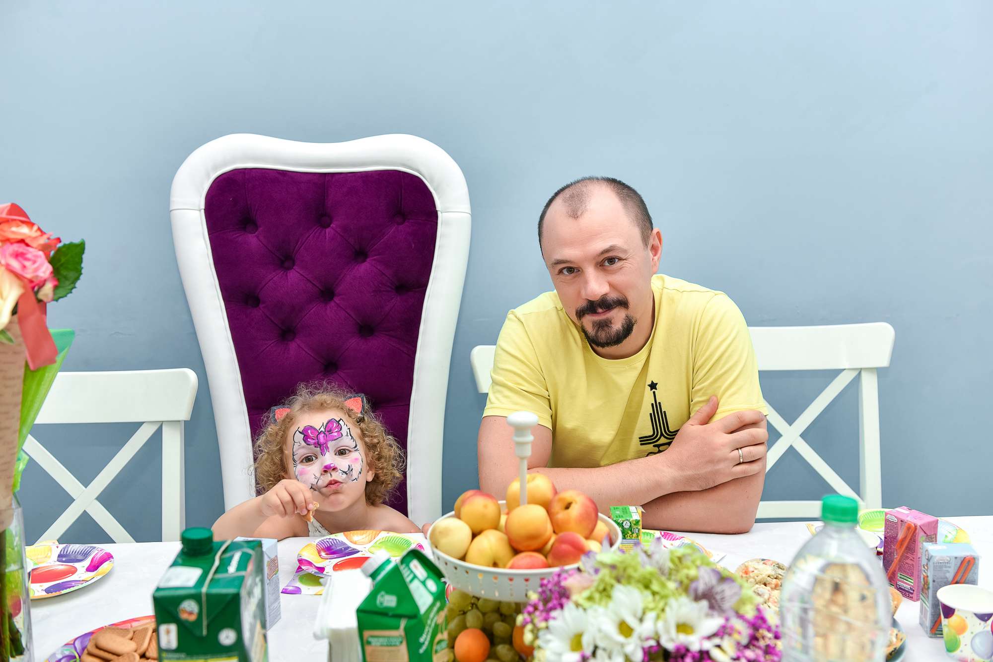 Мужчина в жёлтой футболке и девочка с раскрашенным лицом за столом с фруктами, соками и цветами на праздничной вечеринке.