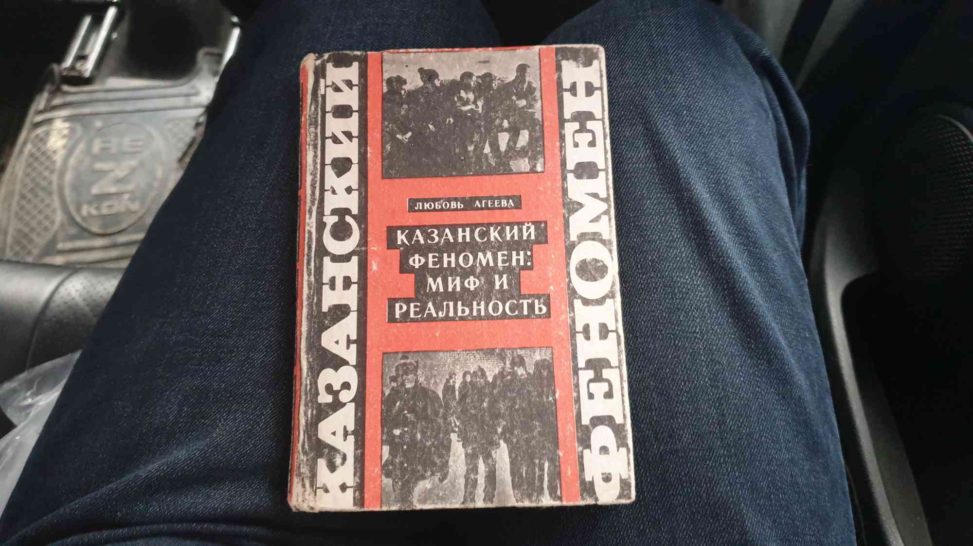 Книга Любови Агеевой «Казанский феномен: миф и реальность» с чёрно-белыми фотографиями на обложке, лежащая на коленях в джинсах внутри автомобиля.