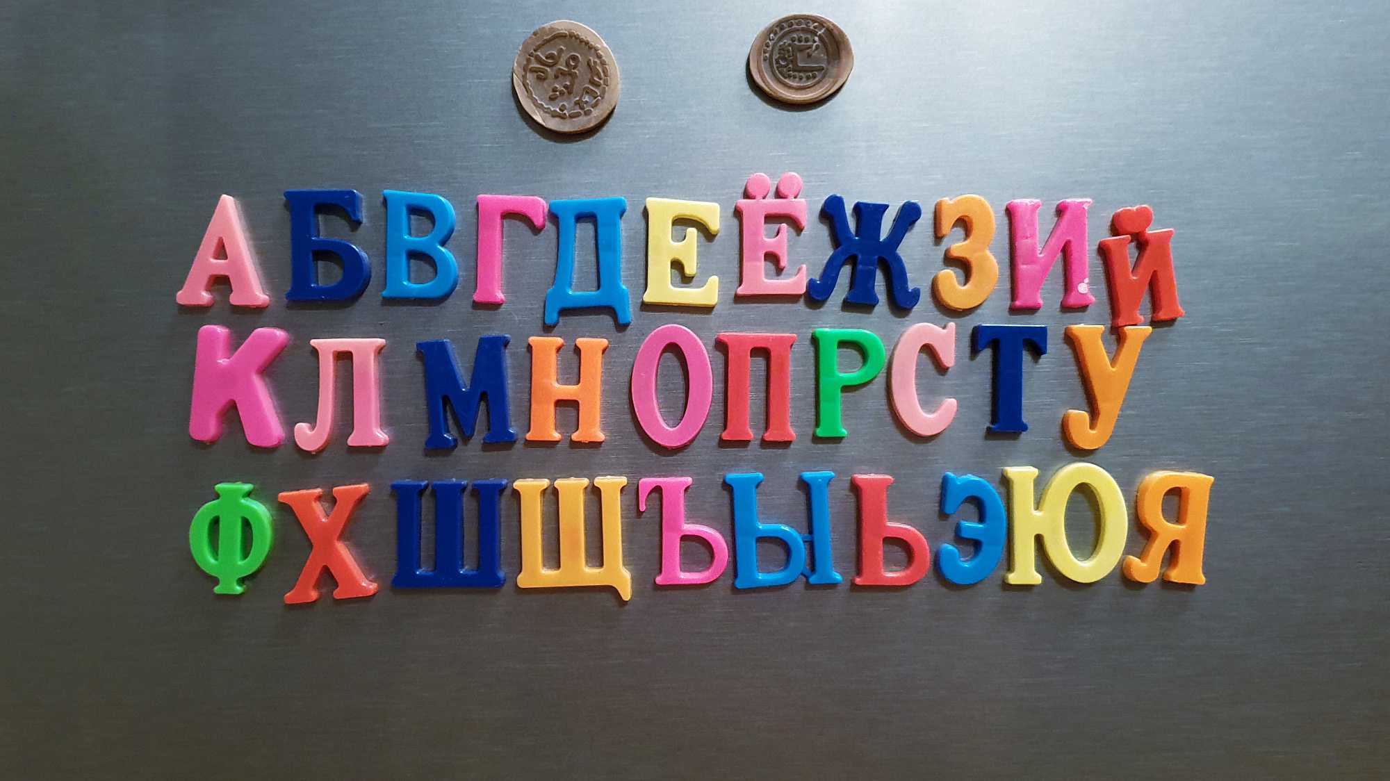Цветные магнитные буквы русского алфавита, выложенные тремя строками на тёмной поверхности, с двумя монетами вверху.